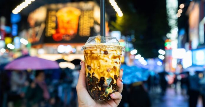Hand holding a caramel-swirled babeltee with tapioca pearls in a lively night market.